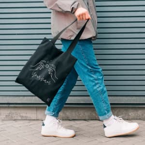 A black tote bag featuring a graphic design, carried by a person wearing blue jeans and white sneakers.