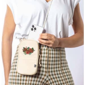 A cream multipurpose bag with floral embroidery and a shoulder strap, featuring a smartphone being held beside it.