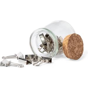 A glass jar with a cork lid containing silver binder clips, some spilling out onto a surface.