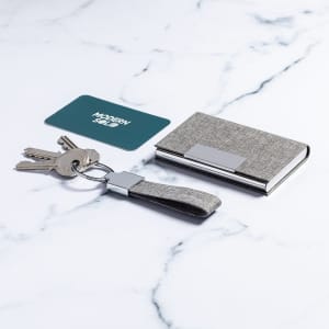 A grey keyring with a strap, keys, and a slim wallet on a marble surface. The wallet has a reflective branding.