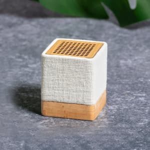 A square, fabric-covered speaker in cream with a wooden base, featuring a mesh top for sound output.