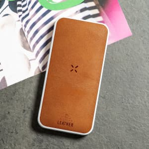 A brown leather power bank with a white border, placed on a textured surface beside colourful magazine pages.