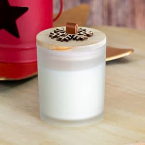 A frosted glass candle with a wooden lid featuring a decorative design on top, placed on a table.