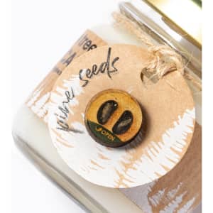 A candle in a glass jar with a gold lid and a tag featuring a wood slice design and a button-shaped embellishment.
