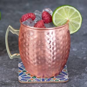A copper mug filled with ice, raspberries, and lime, resting on a decorative coaster.