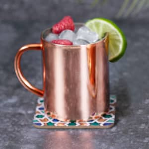 A copper mug filled with ice, raspberries, and a lime wedge, resting on a decorative coaster.