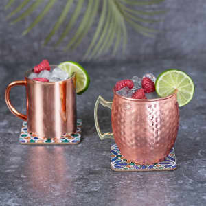 Two coffee mugs in copper with raspberry and lime garnishes, placed on decorative coasters against a grey background.