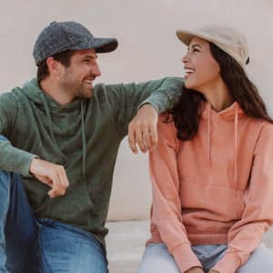 A green cap and a grey cap worn by two people, seated together, smiling, with a neutral background.