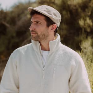 A man wearing a beige Bruck Cap and a white fleece jacket, standing outdoors surrounded by greenery.