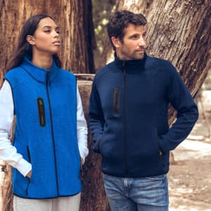 A blue vest and a dark navy vest are displayed on models, set against a backdrop of trees. Each vest features a logo.