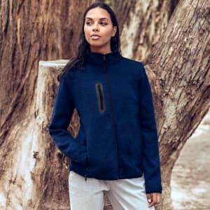 A woman stands next to a tree wearing a navy polar fleece jacket with a zip front and a logo.