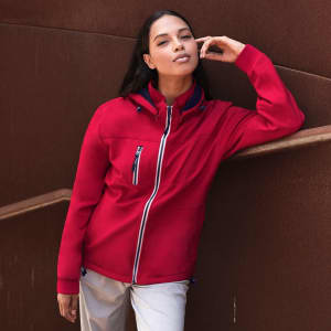 A red soft shell jacket with a hood, featuring zippered pockets and a logo, displayed by a model against a textured background.