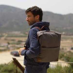 A grey backpack with brown accents, worn by a man outdoors, against a scenic mountainous backdrop.