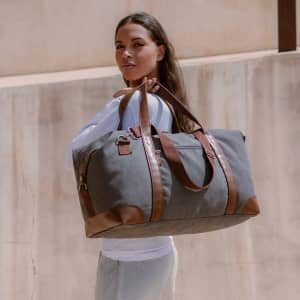 A stylish grey duffel bag with brown leather accents, held by a woman against a textured background.