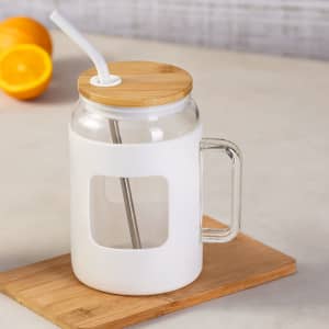 A drink bottle with a glass body, white silicone sleeve, wooden lid, and metal straw on a wooden surface.