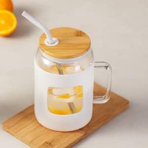 A drink bottle with a clear glass body, bamboo lid, and white sleeve, featuring a straw and a handle.