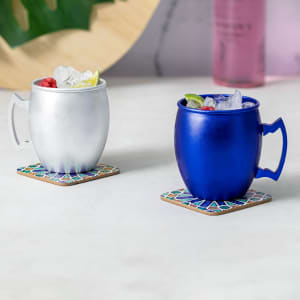A drinkware set featuring a silver mug and a blue mug, both filled with ice and garnished with fruit, on decorative coasters.