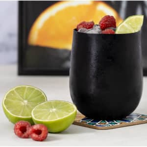 A black plastic cup filled with ice, raspberries, and lime, accompanied by halved limes and a decorative coaster.