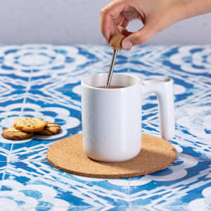 A white coffee mug on a cork mat, with a hand dipping a biscuit into it, beside some cookies.