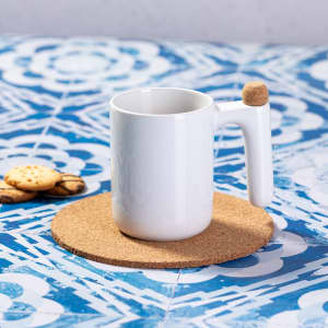 A white coffee mug with a unique handle design, placed on a cork coaster, alongside some cookies.