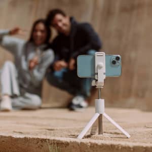 A white tripod holds a smartphone in place, with two people smiling in the background.