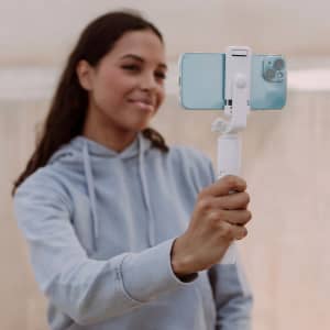 A white tripod holds a smartphone in position, while a smiling woman uses it to take a photo.