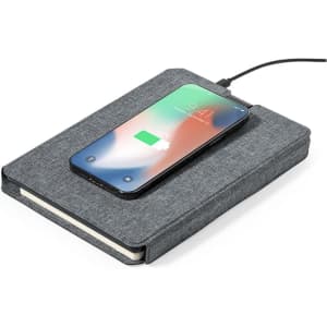 A grey notepad with a textured cover and a smartphone charging on top. The notepad has a logo.