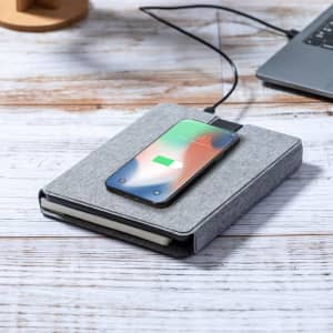 A grey multifunction notepad with a smartphone charging on top, set on a wooden surface beside a laptop.