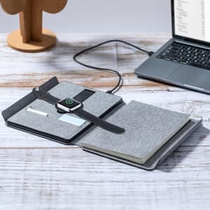 A grey multifunction notepad opens to reveal a notebook, pockets, and a smartwatch charging. A laptop is in the background.