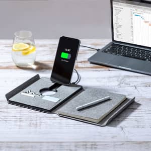 A grey multifunction notepad with a wireless charger, pen, and card holder, set beside a laptop and drink.
