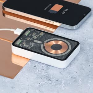 A white Bramson Power Bank with a transparent design sits on a marble surface next to a smartphone and reflective copper objects.