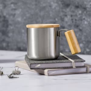 A stainless steel insulated mug with a wooden lid and handle, placed on grey notebooks with binder clips.
