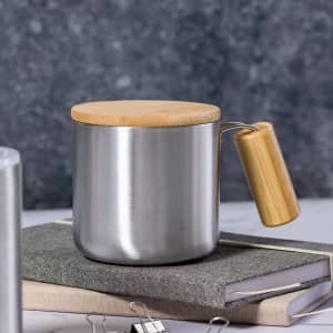 A stainless steel insulated mug with a bamboo lid and a wooden handle, placed on top of a grey notebook and binder clips.