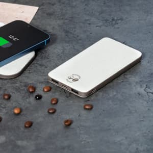 A power bank in light grey sits on a textured surface next to a smartphone, surrounded by coffee beans.