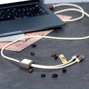 A beige and brown Seymur charging cable lies on a dark surface near a laptop, accompanied by scattered coffee beans.