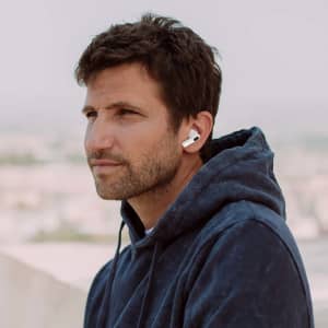 A man wearing white Shima Earphones, dressed in a navy blue hoodie, with a blurred cityscape in the background.