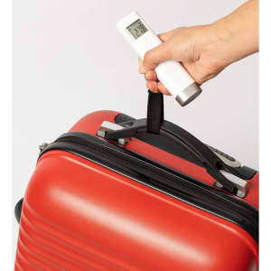 A hand holds a white luggage scale above a red suitcase, measuring its weight with a digital display.