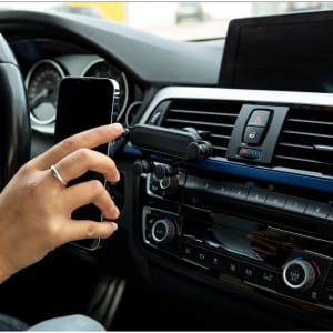 A hand holds a smartphone near a black car phone holder mounted on a dashboard air vent.