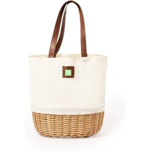 A cream canvas picnic basket with a woven bottom and brown leather handles, featuring a small green square detail.