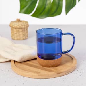 A blue mug with a cork base sits on a wooden tray, accompanied by a cream-coloured cloth and a woven container.