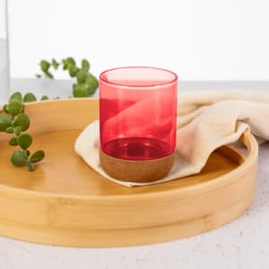 A pink plastic cup with a cork base rests on a neutral cloth within a bamboo tray, surrounded by greenery.