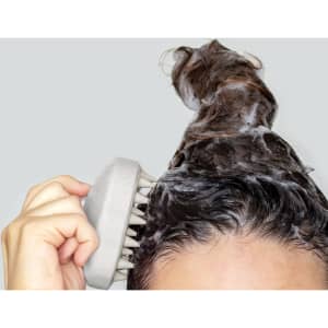 A person using a scalp massager on wet hair, which is lathered with shampoo. The background is grey.