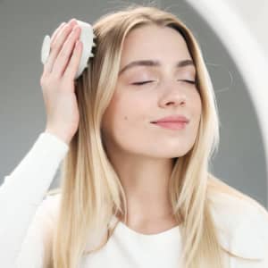 A woman with long blonde hair is gently using a white scalp massager while smiling.