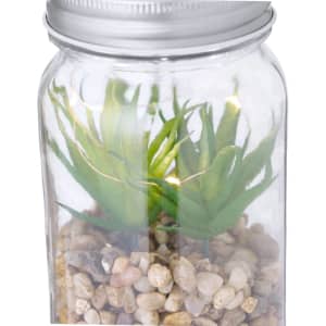 A glass jar containing faux green plants atop a bed of small decorative pebbles, with a metallic lid.