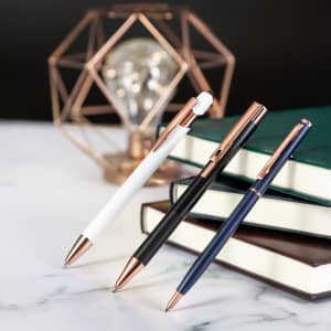 Three stylus touch ball pens in white, black, and navy, with rose gold accents, placed beside stacked notebooks.