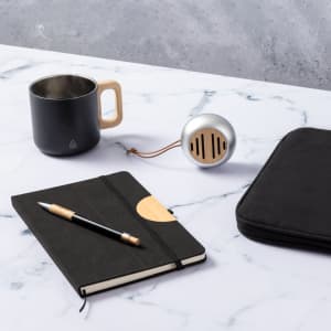A coffee mug with a black finish and wooden handle, placed beside a notebook, pen, and speaker on a marble surface.