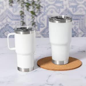 Two white insulated mugs with stainless steel accents on a cork coaster, set against a patterned background.