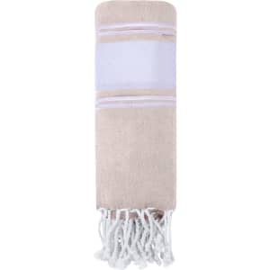 A beige Lainen Towel Pareo with white stripes and tassels at the ends, neatly rolled up. It features a branding logo.