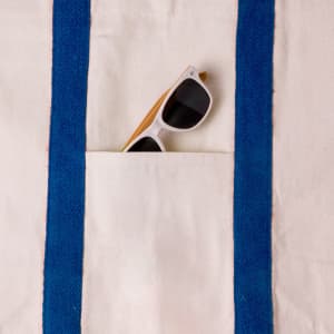 A tote bag with blue straps and a front pocket featuring a pair of white sunglasses partially tucked inside.