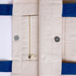 A beige tote bag with blue handles featuring a zip pocket and snap closures. It has a simple, minimalist design.